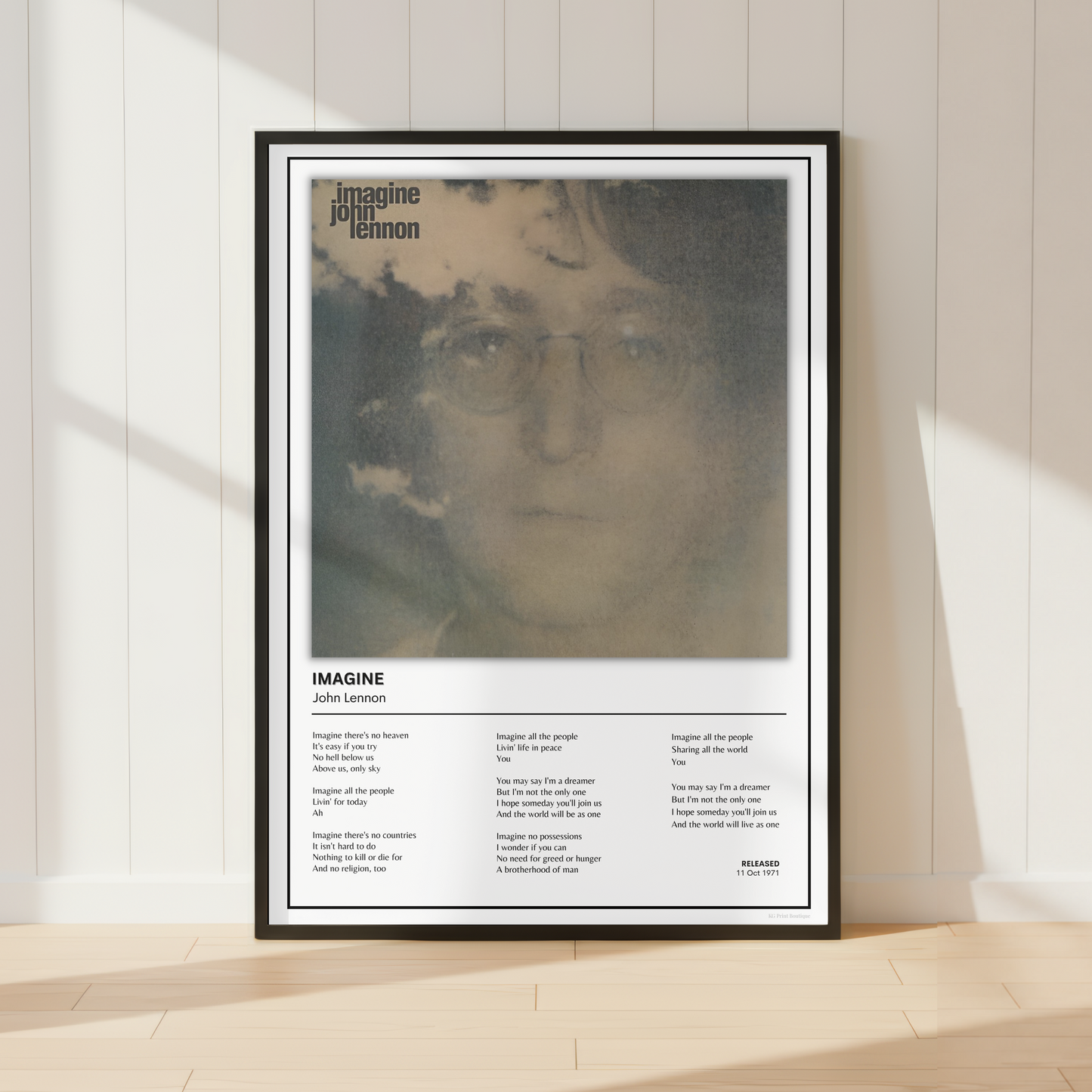 A framed John Lennon 'Imagine' poster leaning against a white paneled wall on a light wood floor, with soft sunlight casting shadows across the scene.