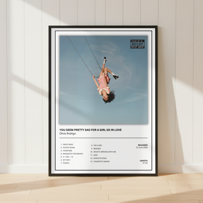 A framed music poster of Olivia Rodrigo's song 'You See Pretty Sad For A Girl So In Love' leaning against a white paneled wall on a light wood floor.