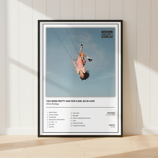 A framed music poster of Olivia Rodrigo's song 'You See Pretty Sad For A Girl So In Love' leaning against a white paneled wall on a light wood floor.