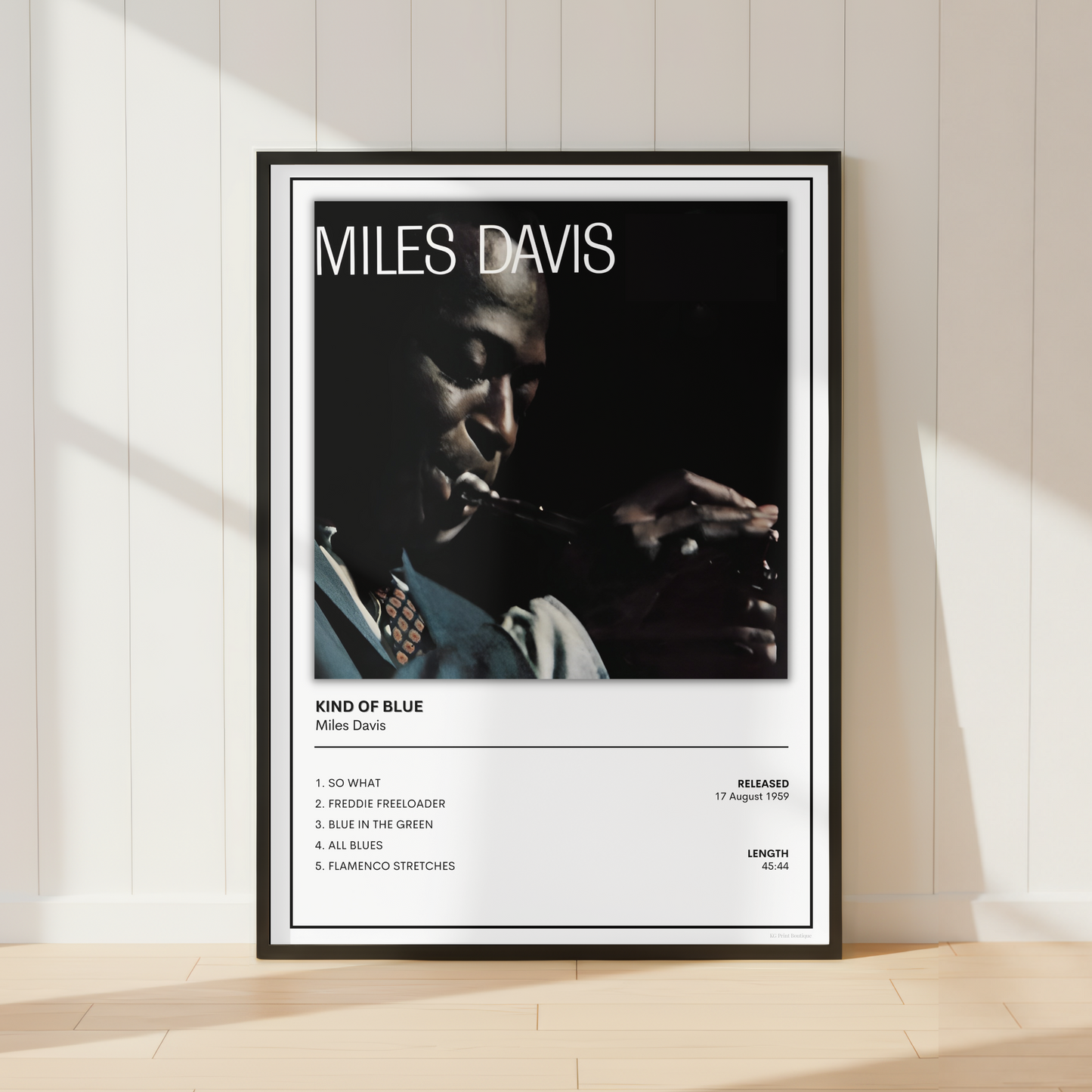 A framed Miles Davis 'Kind of Blue' music poster leaning against a white paneled wall on a light wood floor, with soft sunlight casting shadows across the scene.