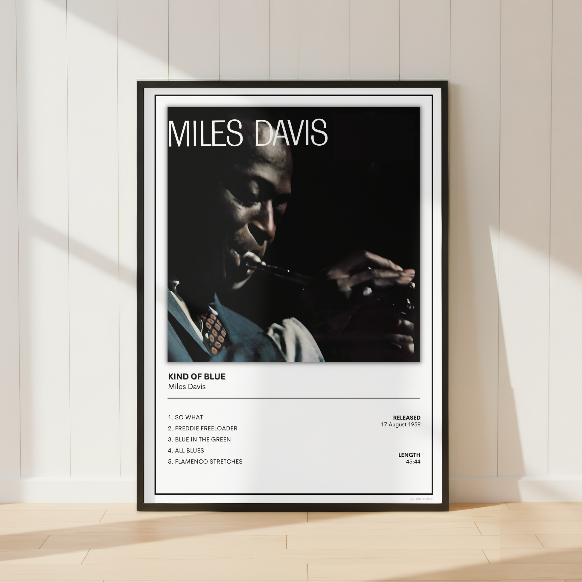 A framed Miles Davis 'Kind of Blue' music poster leaning against a white paneled wall on a light wood floor, with soft sunlight casting shadows across the scene.