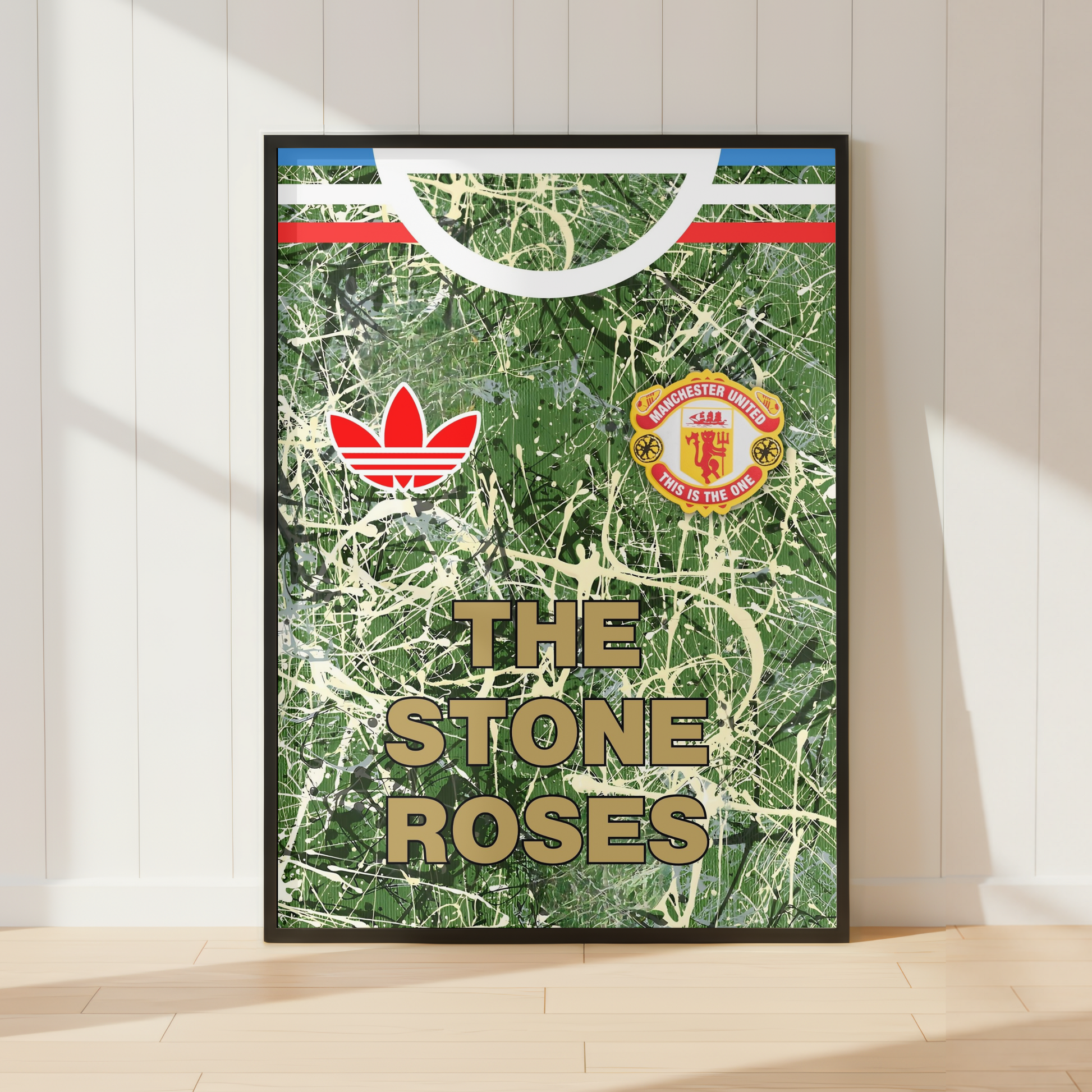 A framed art print of a Manchester United football jersey design, leaning against a white paneled wall on a light wood floor. The print features a green marble pattern with white veins, the red Adidas logo, the Manchester United crest, and the text 'THE STONE ROSES'.