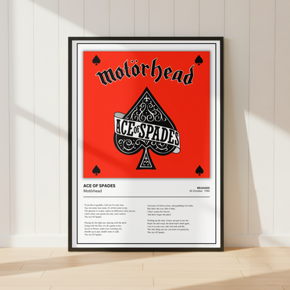 A framed Motorhead 'Ace of Spades' poster leaning against a white paneled wall on a light wood floor, with soft diagonal shadows across the scene.