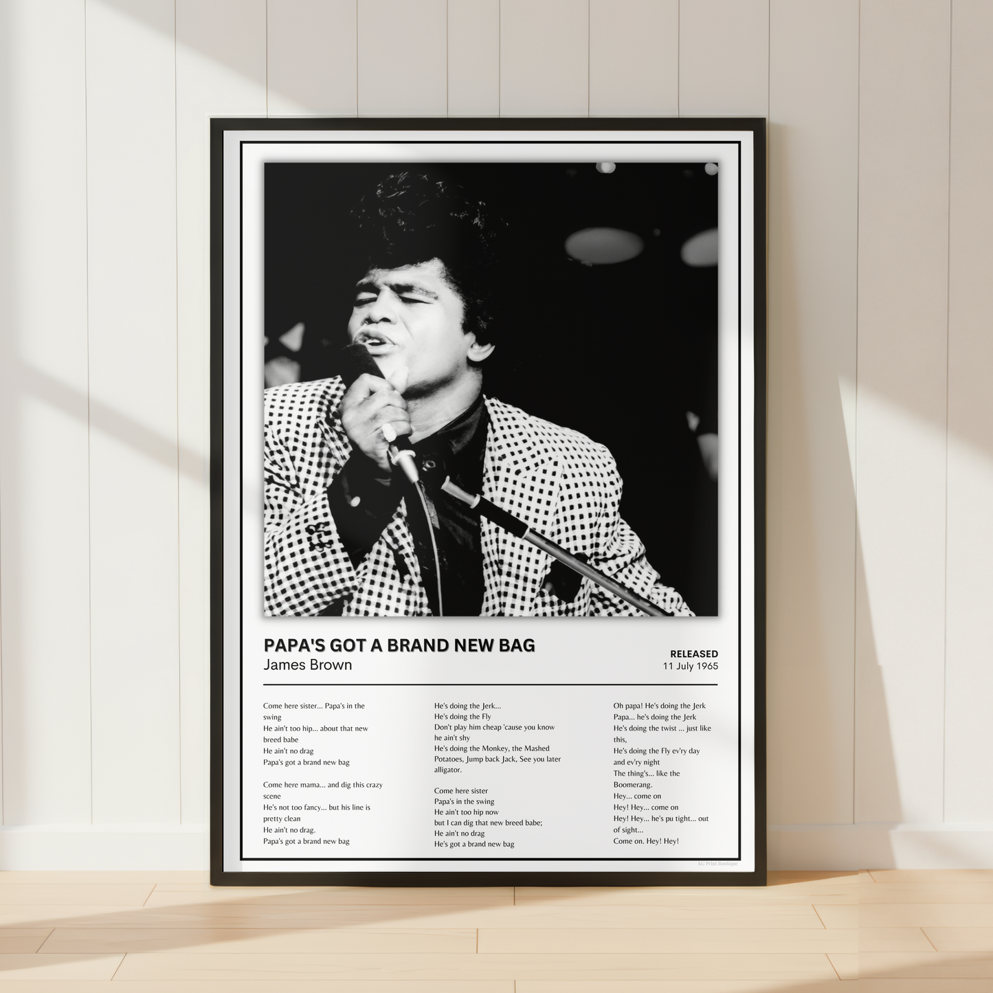 A framed black-and-white music poster of James Brown leaning against a white paneled wall on a light wood floor. The poster features a portrait of James Brown singing into a microphone, with the text 'PAPA'S GOT A BRAND NEW BAG' and a tracklist below.