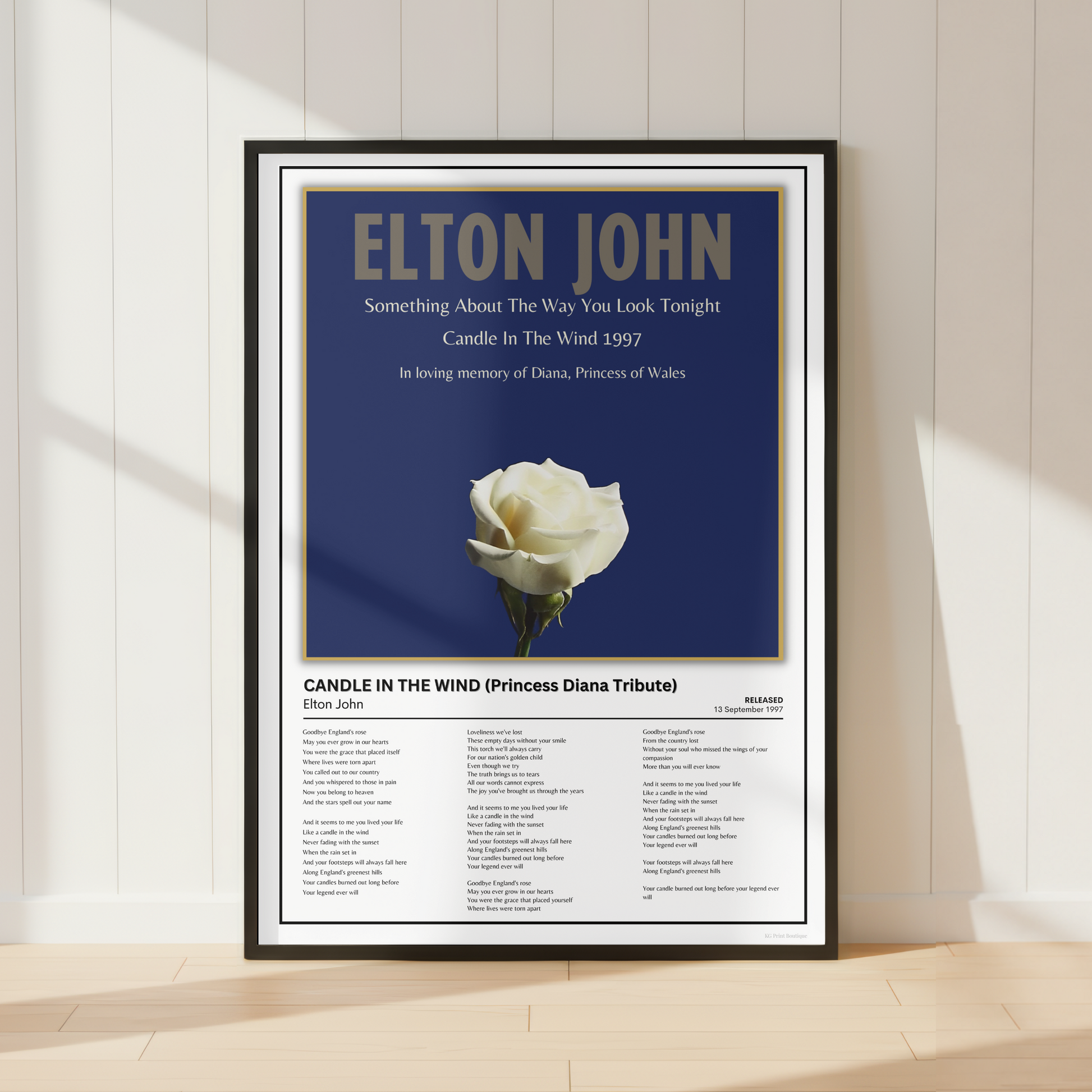A framed Elton John tribute poster leaning against a white paneled wall on a light wood floor. The poster features a dark blue background with the text 'ELTON JOHN' and a single white rose, plus song lyrics at the bottom.