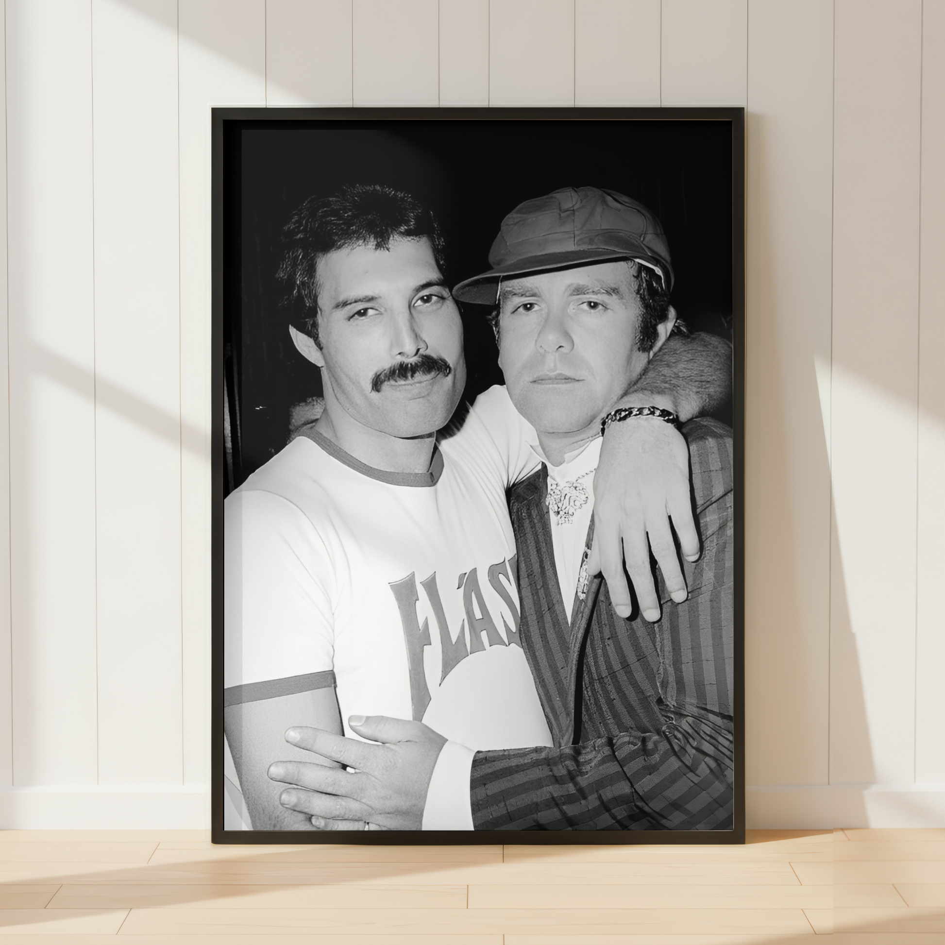 A black-framed black and white photographic print of Freddie & Elton leaning against a white paneled wall on a light wood floor, with soft sunlight casting shadows across the wall and floor.