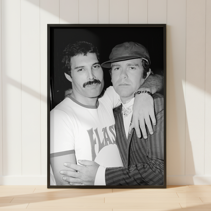 A black-framed black and white photographic print of Freddie & Elton leaning against a white paneled wall on a light wood floor, with soft sunlight casting shadows across the wall and floor.