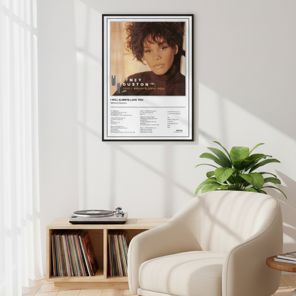 A framed Whitney Houston 'I Will Always Love You' music poster is displayed leaning against a white paneled wall on a light wood floor, with soft sunlight casting shadows across the scene.