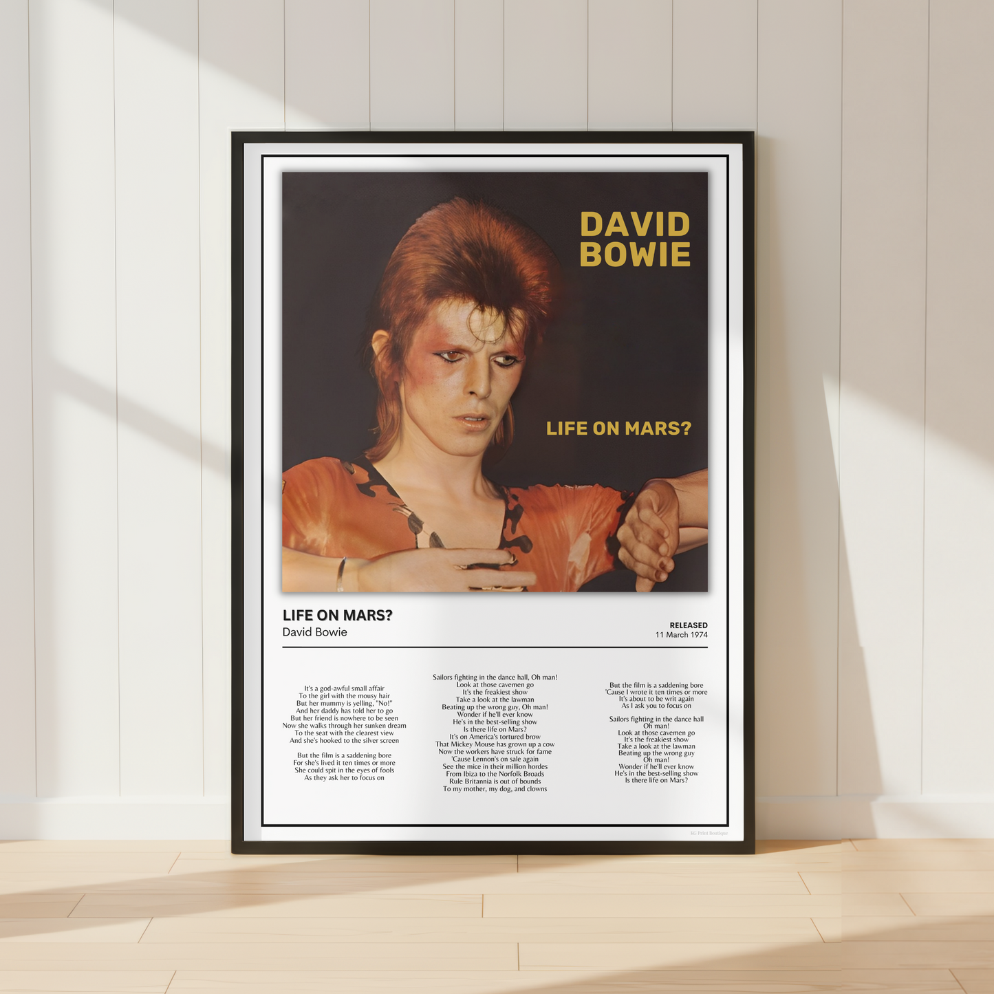 A framed David Bowie 'Life on Mars?' poster leaning against a white paneled wall on a light wood floor, with soft window shadows cast across the scene.