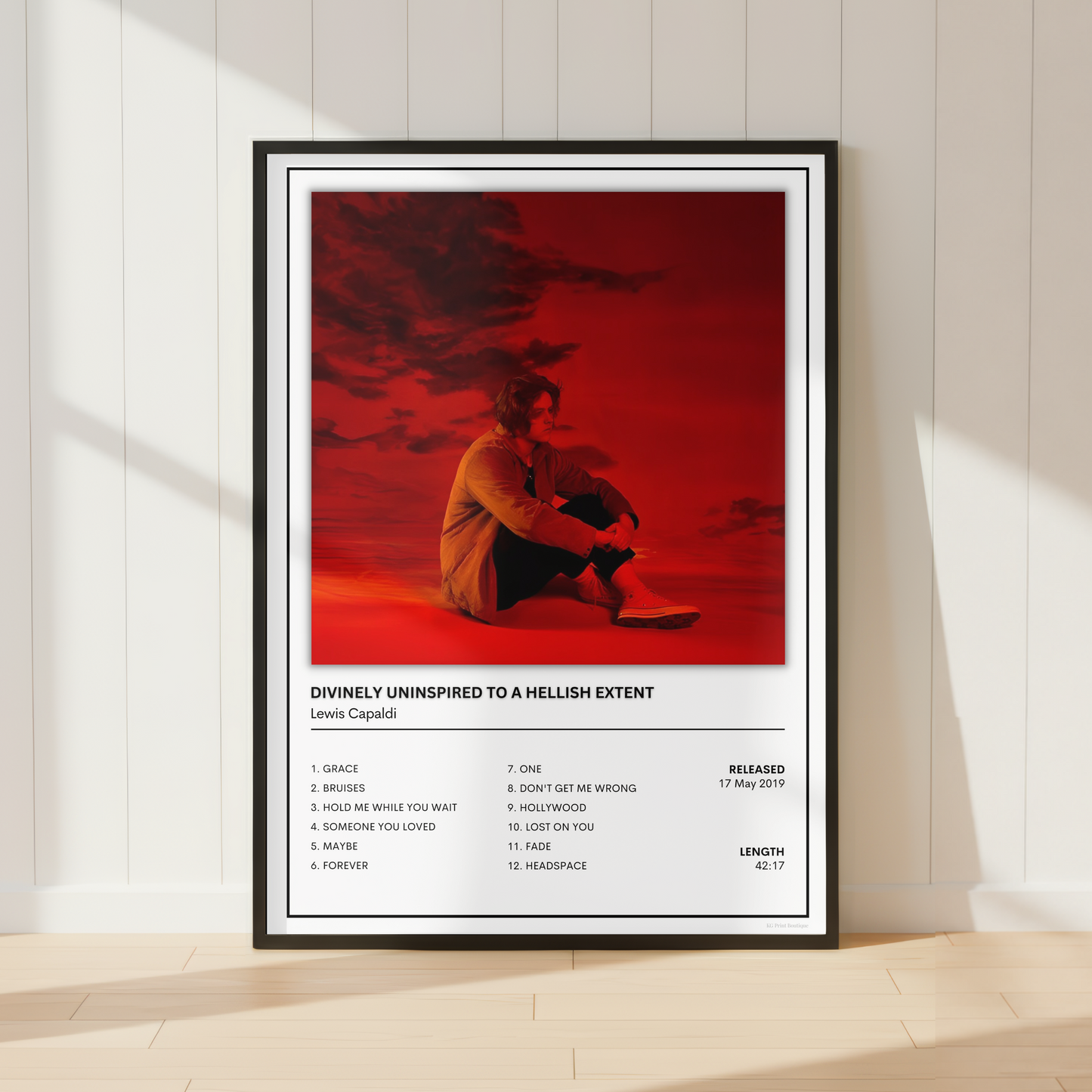 A framed music poster for Lewis Capaldi's 'Divinely Uninspired to a Hellish Extent' leaning against a white paneled wall on a light wood floor, with warm sunlight casting shadows.