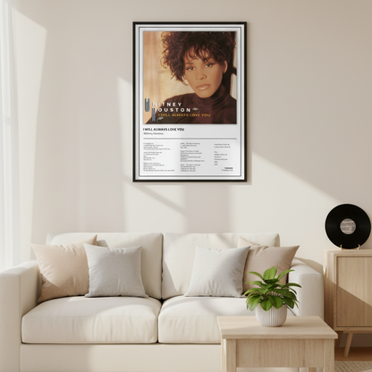 A framed Whitney Houston 'I Will Always Love You' music poster is displayed leaning against a white paneled wall on a light wood floor, with soft sunlight casting shadows across the scene.