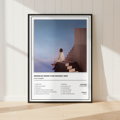 A framed music poster of Lewis Capelli's 'Broken By Desire To Be Heavily Sent' leaning against a white paneled wall on a light wood floor.