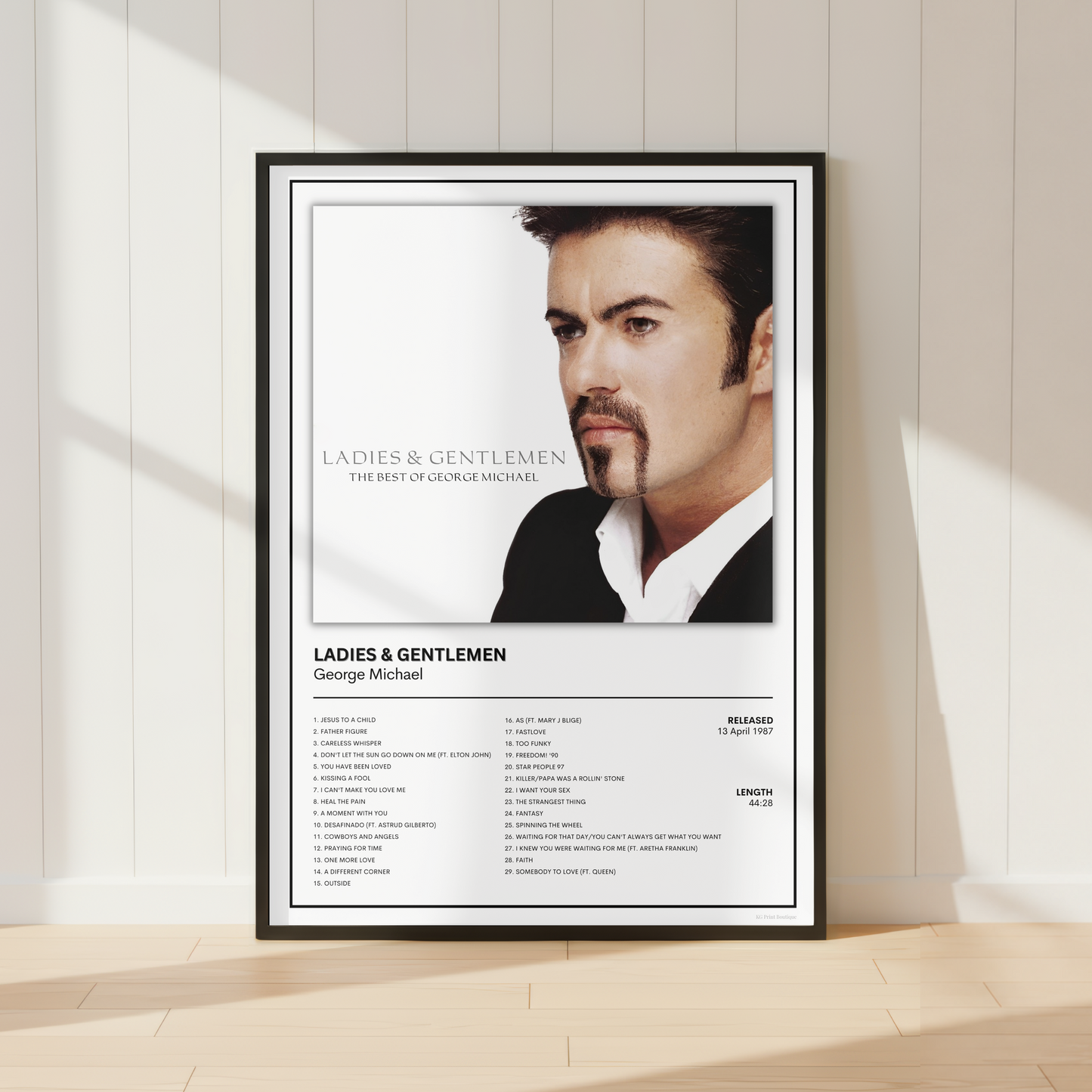 A framed music poster of George Michael's album 'Ladies & Gentlemen' leaning against a white paneled wall on a light wood floor, with soft sunlight casting shadows.