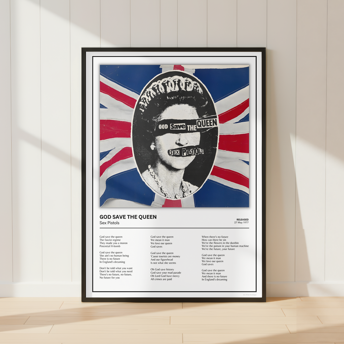 A framed music poster of Queen's 'God Save the Queen' leaning against a white paneled wall on a light wood floor, featuring a stylized portrait over a Union Jack background.