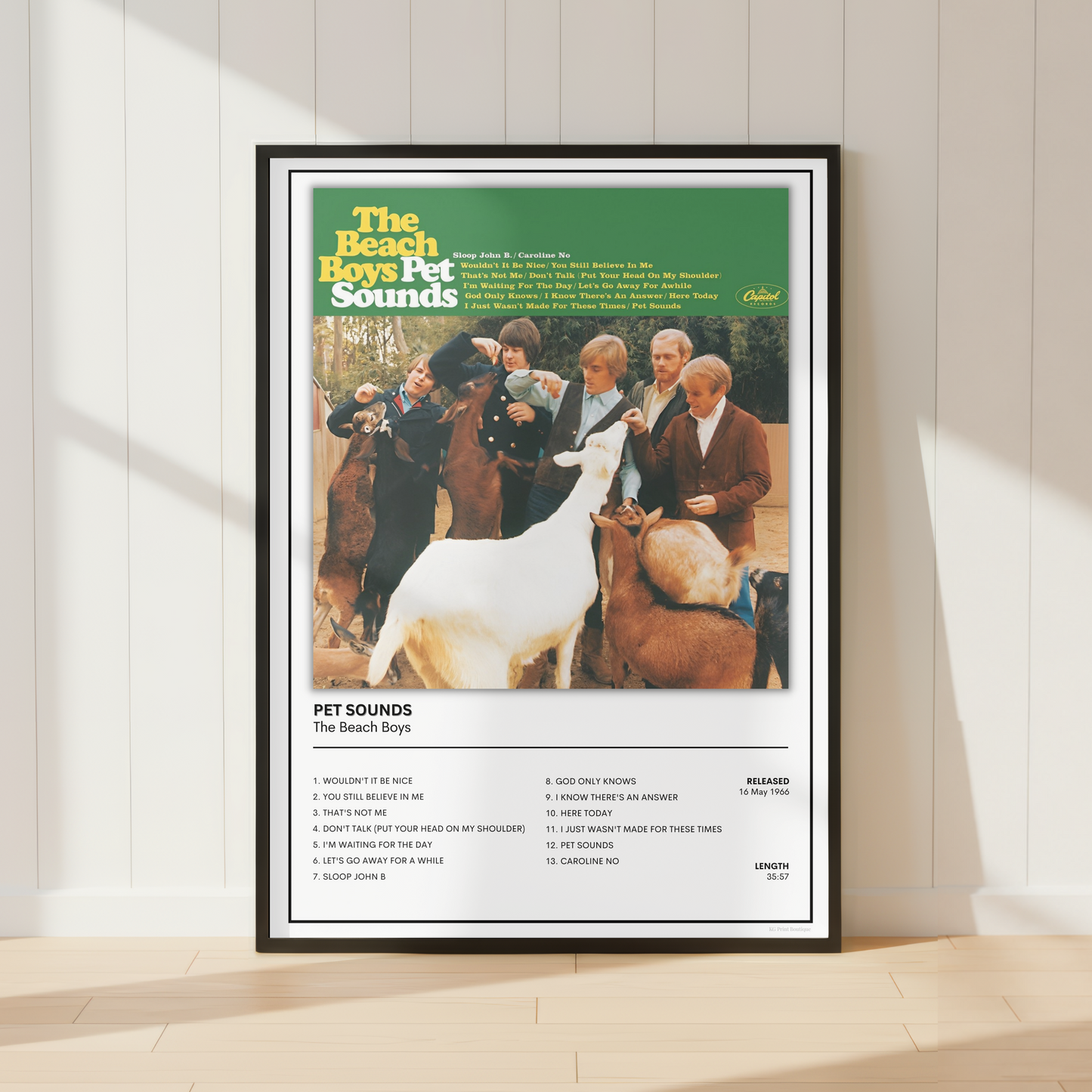 A framed retro-style poster for 'The Beach Boys Pet Sounds' stands against a white paneled wall on a light wood floor. The poster features the album cover artwork with the title at the top, a central photo of people and pets, and a tracklist at the bottom.