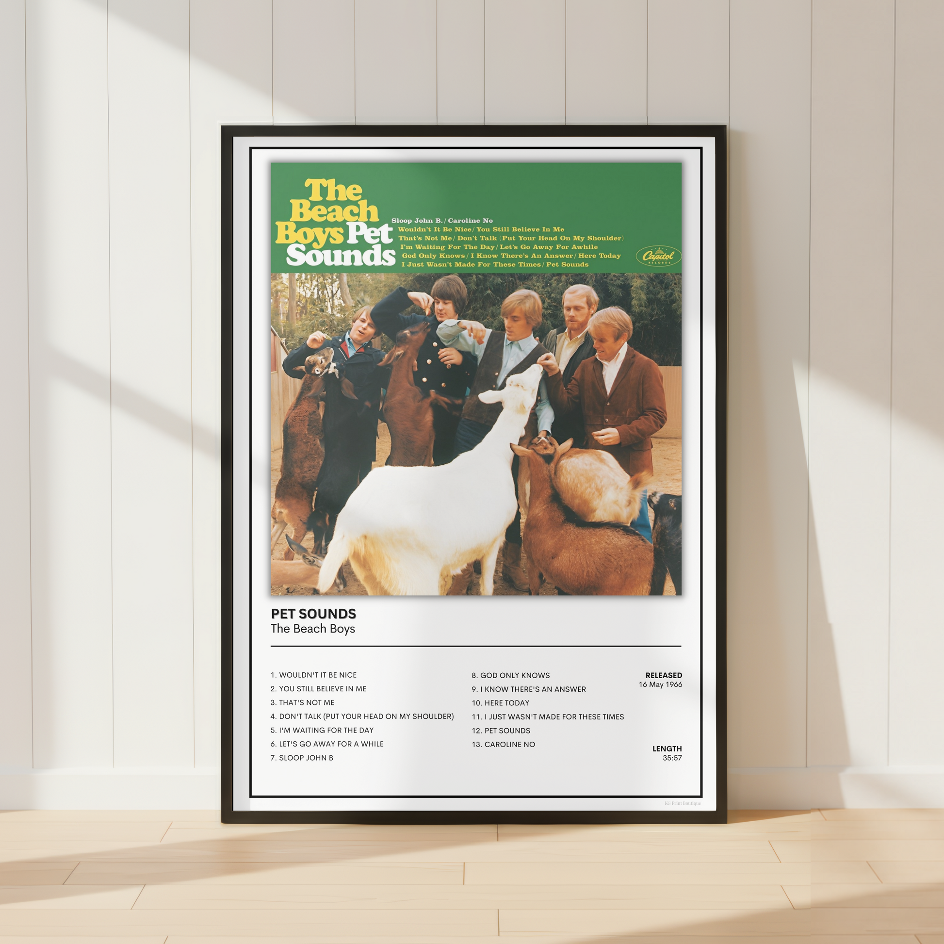 A framed retro-style poster for 'The Beach Boys Pet Sounds' stands against a white paneled wall on a light wood floor. The poster features the album cover artwork with the title at the top, a central photo of people and pets, and a tracklist at the bottom.