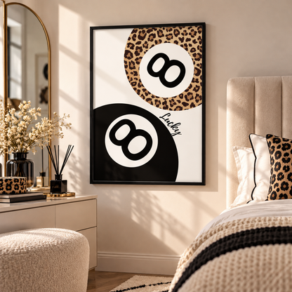 A framed wall art print featuring two black-and-white 8-ball motifs with one leopard-print circle, displayed in a styled bedroom above a bed with matching leopard-print pillows.