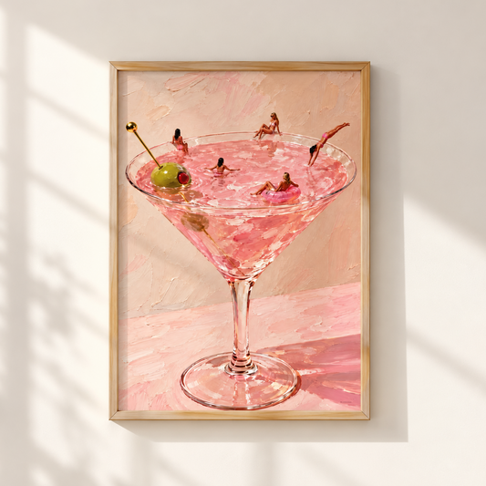A framed art print depicting a pink martini cocktail in a martini glass with a green olive on a pink background, hung on a white wall with soft window shadows.