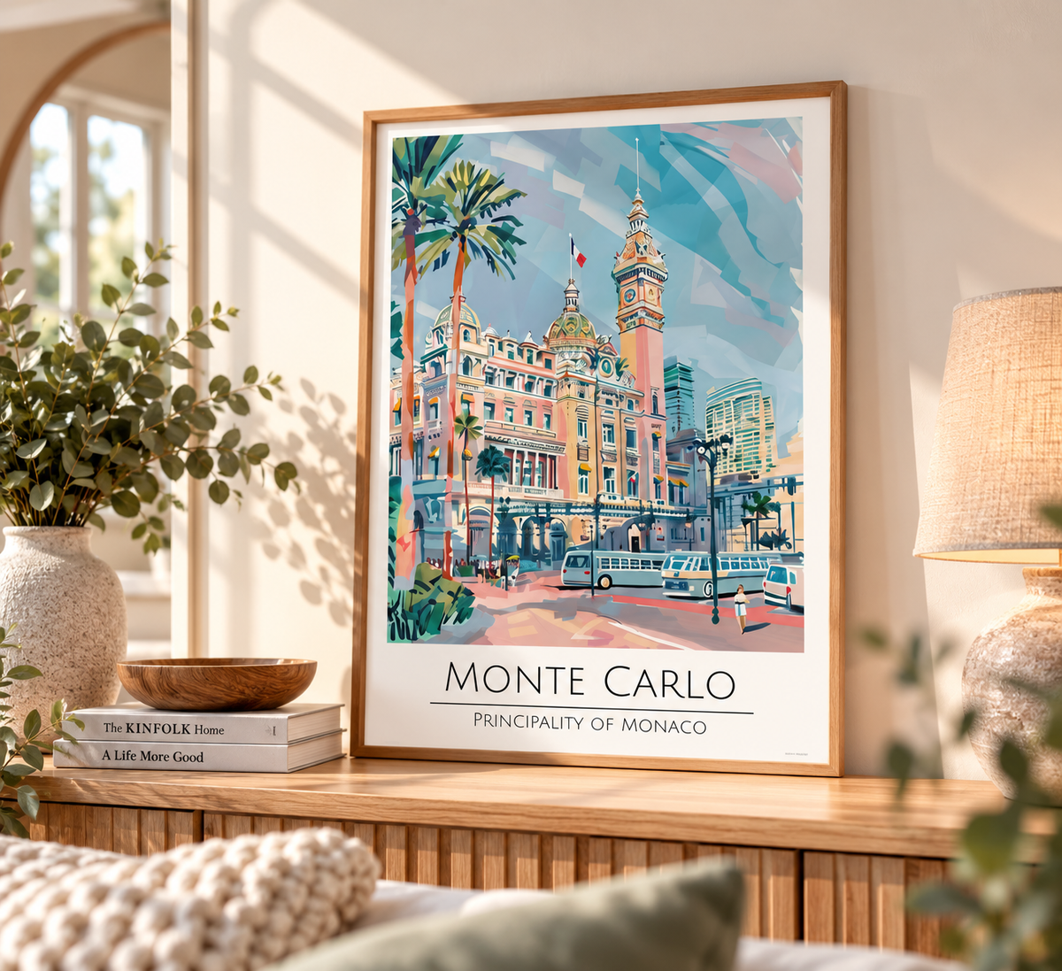 A framed travel poster of Monte Carlo, Principality of Monaco, displayed on a wooden console table in a cozy, sunlit room with plants and a lamp.