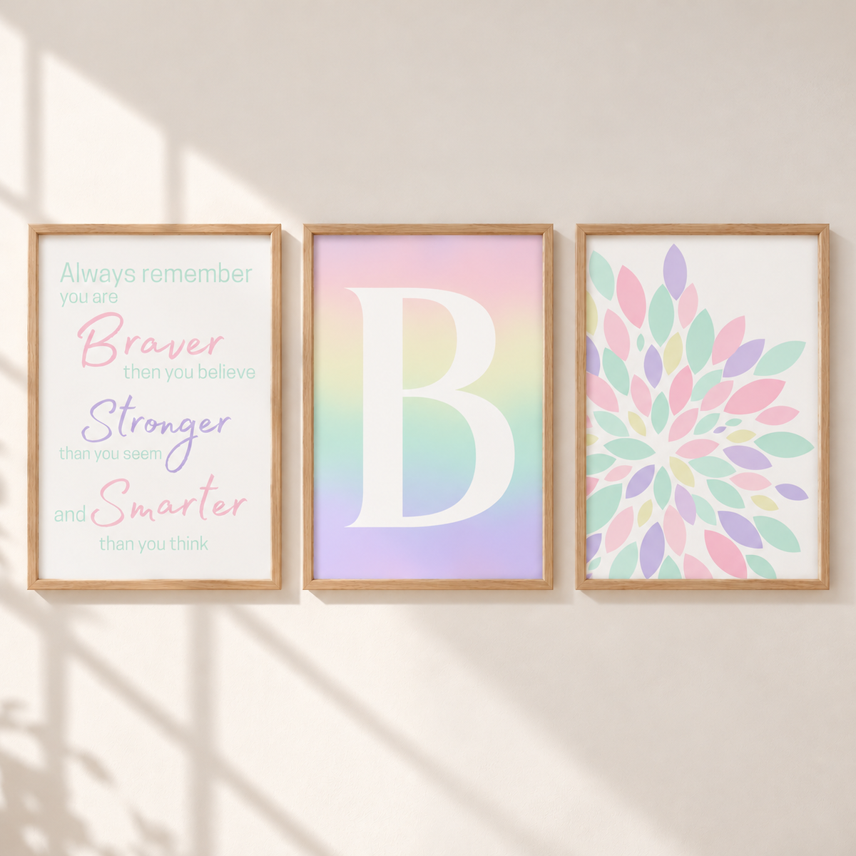 Three framed wall art prints displayed side by side on a warm beige wall with soft shadow overlays. The left print features motivational text in pastel colors, the center print shows a large white letter B over a pastel rainbow gradient, and the right print displays a colorful floral mandala pattern.