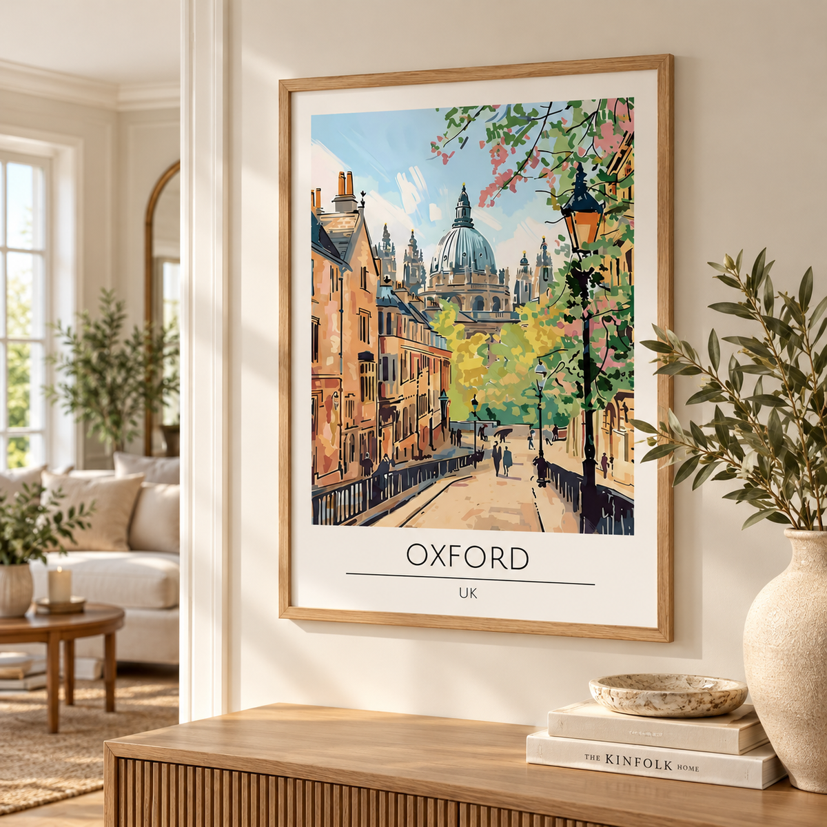 A framed art print of an Oxford street scene hangs on a beige wall in a styled living room. The print features a colorful illustration of Oxford landmarks, including a domed cathedral and trees with pink and yellow leaves, with 'OXFORD UK' printed at the bottom.