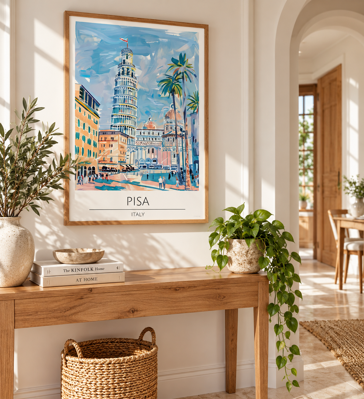 A framed art print of Pisa, Italy, featuring a colorful painting of the Leaning Tower of Pisa and palm trees, styled on a wooden console table in a bright, modern room with plants.