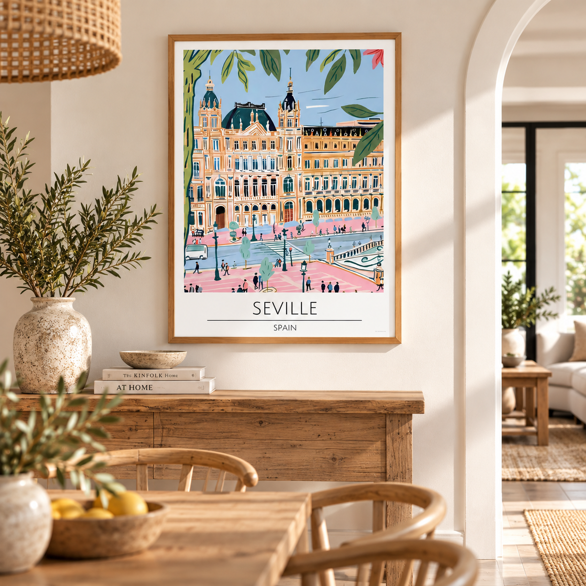A framed travel poster of Seville, Spain, featuring a colorful illustration of the historic Plaza de España building, displayed on a wooden console table in a warm, modern dining room setting.