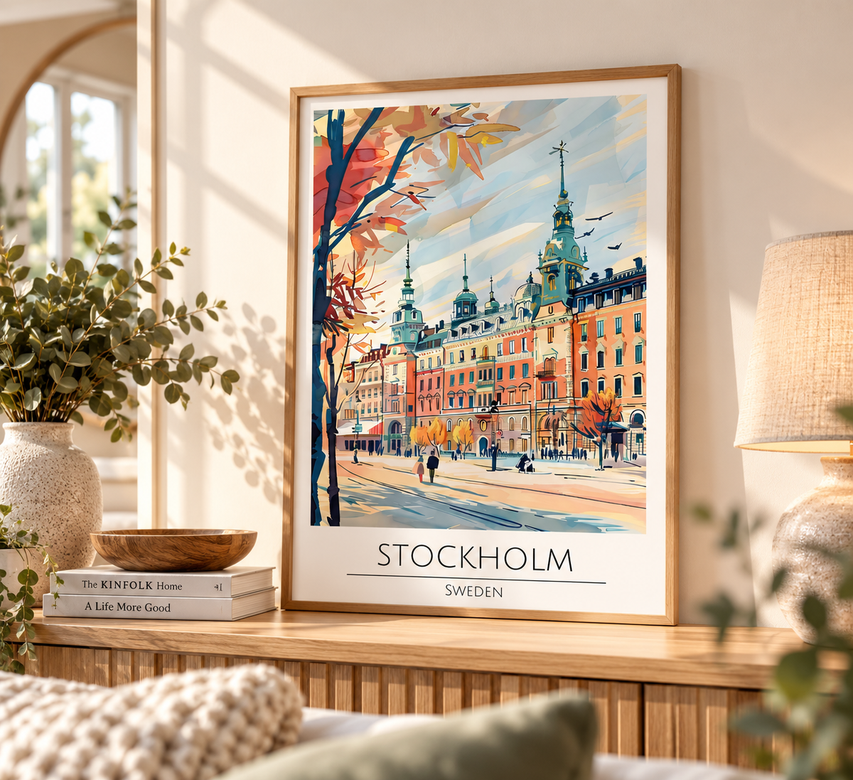 A framed travel poster of Stockholm, Sweden, displayed on a wooden console table in a cozy, sunlit room. The poster features a colorful watercolor illustration of the city's architecture and autumn trees, with 'STOCKHOLM SWEDEN' printed at the bottom.