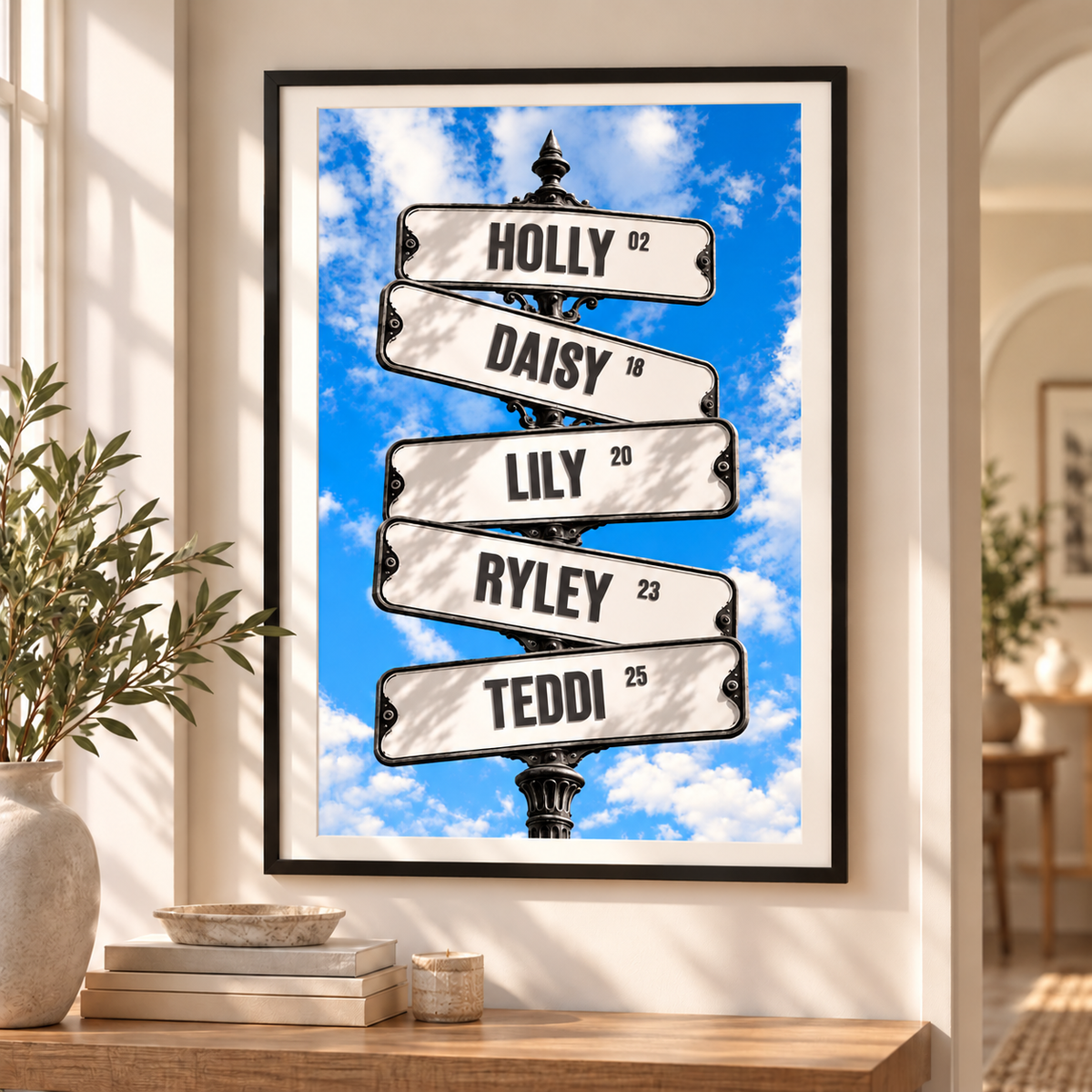 A framed personalized street sign art print hangs on a beige wall above a wooden console table. The print features five white curved street signs on a black pole against a bright blue sky with white clouds, with names 'HOLLY', 'DAISY', 'LILY', 'RYLEY', and 'TEDDI' and ages printed on them.