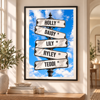 A framed personalized street sign art print hangs on a beige wall above a wooden console table. The print features five white curved street signs on a black pole against a bright blue sky with white clouds, with names 'HOLLY', 'DAISY', 'LILY', 'RYLEY', and 'TEDDI' and ages printed on them.