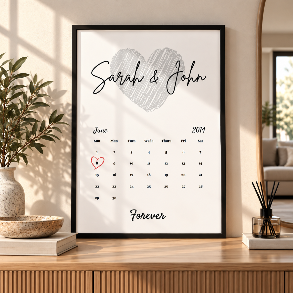 A framed personalized calendar print with a large heart and the names 'Sarah & John' displayed on a light wood console table in a styled home interior.